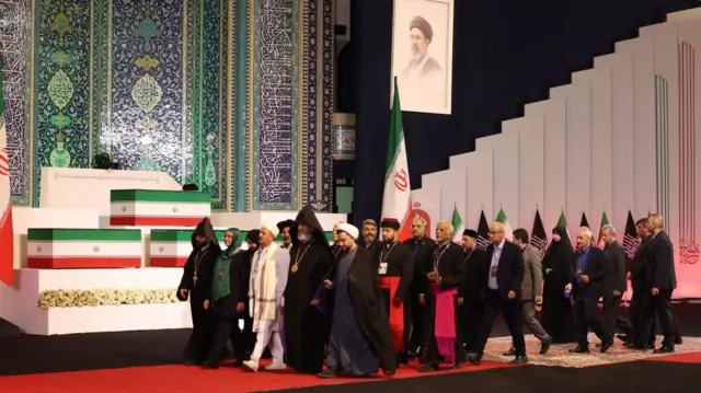 ハメネイ前最高指導者の遺影が掲げられ、白い壇上にイラン国旗で包まれた棺が並ぶ前を、さまざまな宗教の聖職者やスーツ姿の人たちが通る