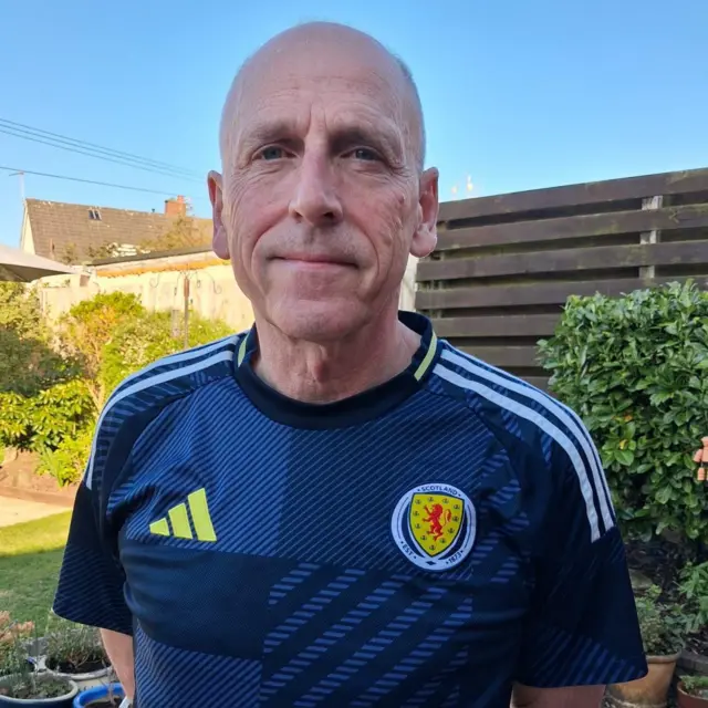 Hamish Husband de pé no seu jardim, vestindo uma camisa da seleção escocesa de futebol