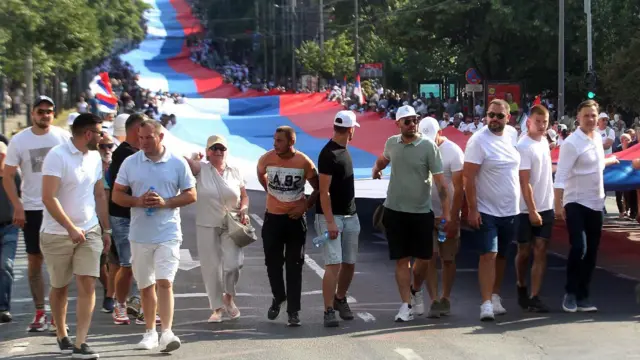Људи ходају улицом, иза њих застава Србије