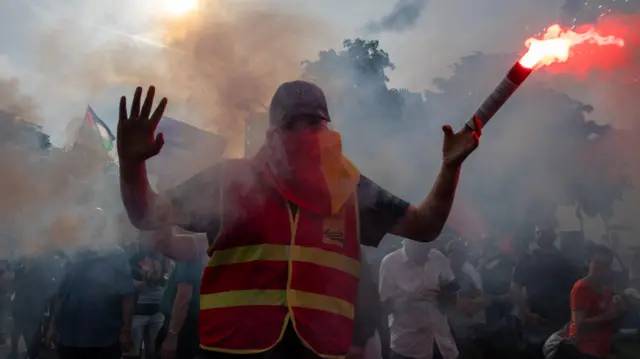 Un homme vêtu d'une veste haute visibilité et portant des lunettes de soleil agite une fusée rouge allumée lors d'une manifestation en France.