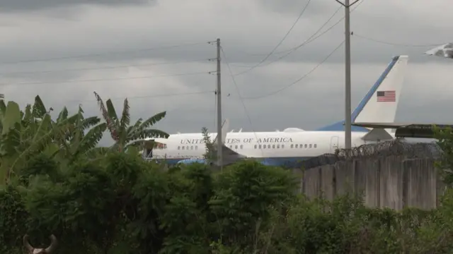 Un avión blanco con letras azules que dicen "Estados Unidos de América", rodeado por vegetación. También se ve un muro con un alambrado de púas.