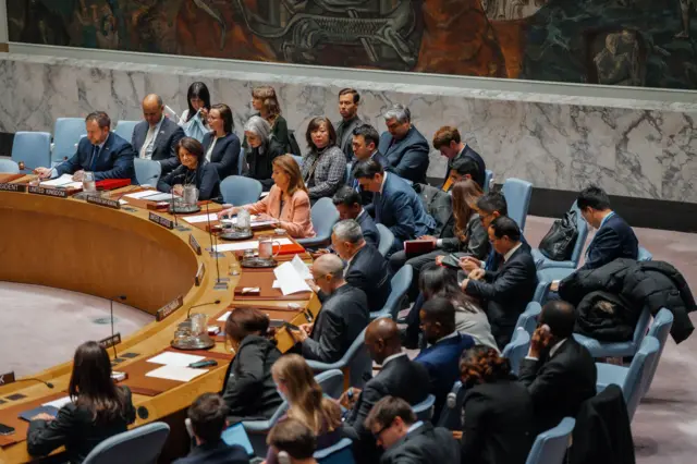 Засідання у великій залі ООН: делегації сидять за круглим столом, перед ними – документи, мікрофони та таблички з назвами країн. Інтер’єр прикрашений мармуровими стінами та великим настінним мистецьким панно.