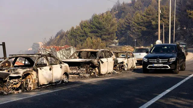 Кілька повністю вигорілих автомобілів стоять уздовж дороги після лісової пожежі. На тлі видно обпалений лісовий схил, а повз пошкоджені машини рухаються вцілілі автомобілі.