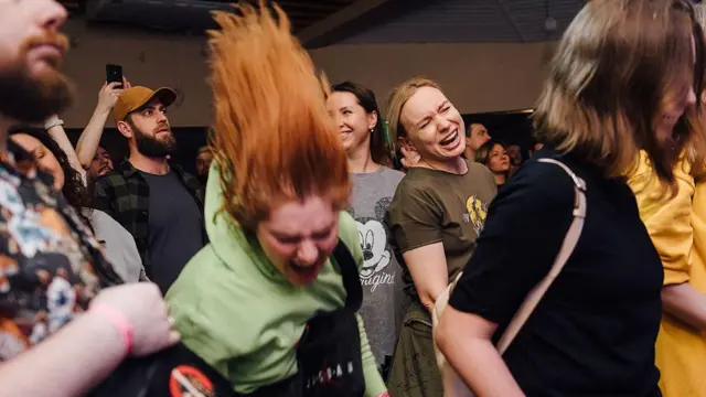 Люди в різному вбранні слемляться під музику, у всіх різні емоції на обличчі, та особливо виділяється рудоволоса дівчина в худі