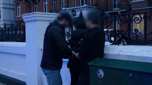 На фото пресс-службы полиции Лондона двое полицейских с заблюренными лицами задерживают человека возде ограды с белым постаментом и узорными решетками. Фото сделано в вечернее время - в сумерках