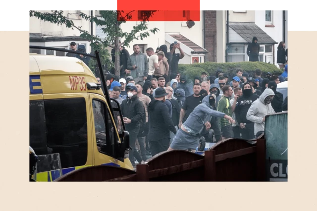 A crowd of male protesters behind a police van. One man in the foreground is wearing a black face covering and blue tracksuit top and shorts. He appears to be hurling something.