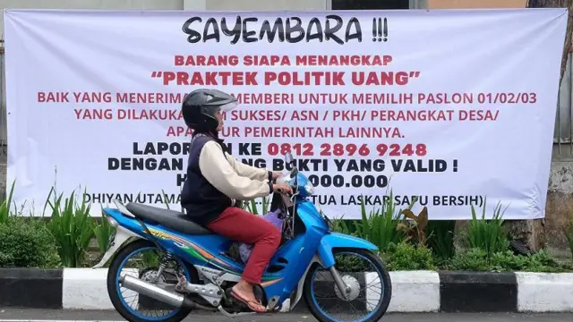 Pengendara sepeda motor melintas di depan spanduk tolak politik uang pada Pilkada 2024 di Temanggung, Jawa Tengah, Senin (25/11/2024).