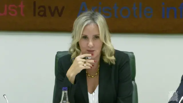 肩まで伸びた金髪の女性が黒いジャケットと白いインナーを着て、マイクの置かれた机に向かって座り、正面を見つめながらペンを持った右手にあごを載せている