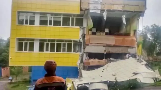 地震で一部が倒壊した建物の前に、救助隊員が立っている