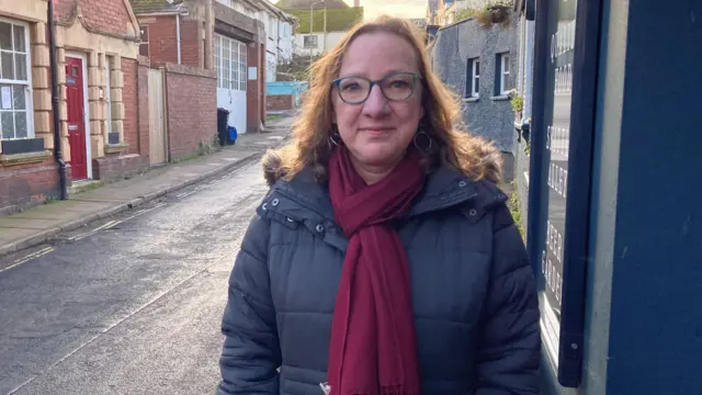 Kerry Wright está de pie junto a un edificio azul a la derecha. Lleva un abrigo gris, una bufanda burdeos y gafas. Tiene el pelo castaño hasta los hombros. Lleva pendientes de aro. Está de pie junto a una carretera. Hay edificios de ladrillo a la izquierda.