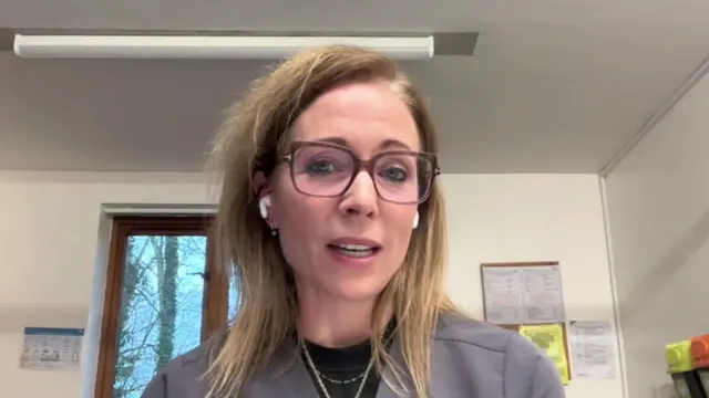 Una mujer de cabello largo y rubio está sentada en la consulta de un médico. Lleva gafas. Habla a la cámara y lleva auriculares.