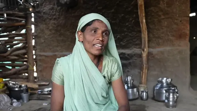 Sushila's mother Shantibai wearing a green traditional Indian saree covering her head sitting in her kitchen. In the background are steel utensils and a logs of wood. 