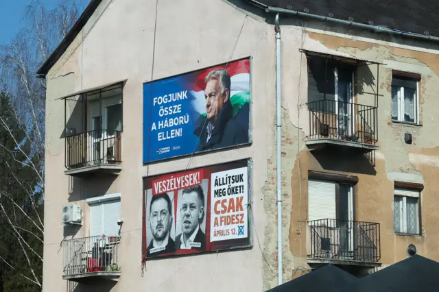 Два плаката на стене дома. Плакат сверху призывает «Давайте объединимся против войны», а нижний, с портретами Мадьяра и Зеленского, предупреждает: «Они опасны! Остановим их! Только Фидес. 12 апреля»