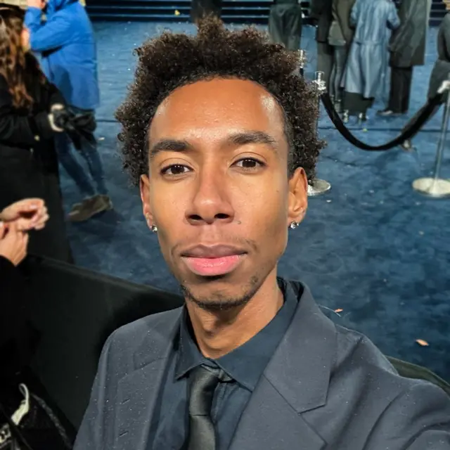 Un joven elegantemente vestido posa para una selfi frente a una alfombra azul. Se ven las cuerdas de terciopelo de las zonas VIP y la gente deambulando esperando la llegada de invitados importantes.