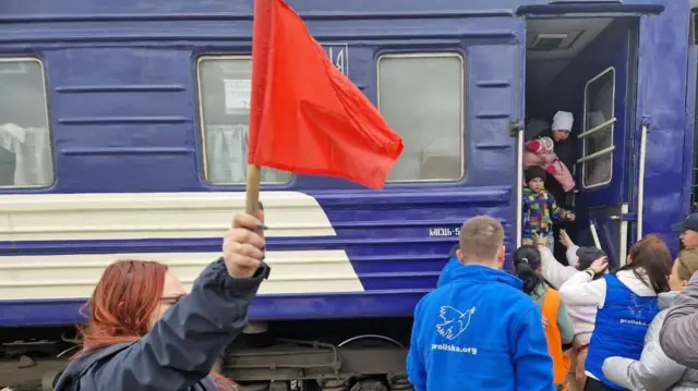 Жінка у синій формі тримає червоний прапорець біля потяга. З вагона виходять люди.
