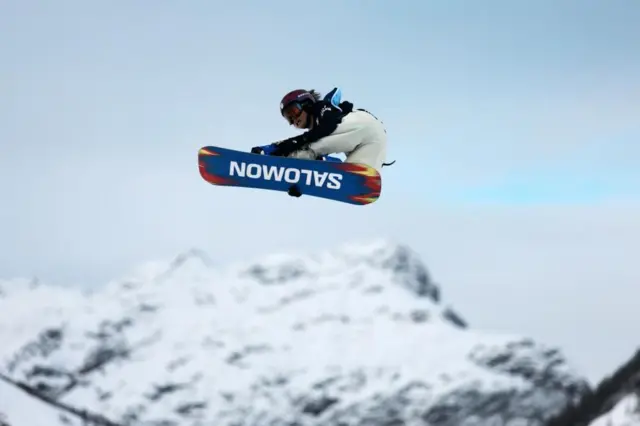 黒っぽいヘルメットとゴーグル、白いパンツを着けた村瀬が空中でバランスをとっている。背景には雪をかぶった山脈がぼんやり見える