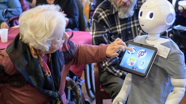 
Uma mulher idosa olha para o robô e toca a tela em sua parte frontal.
