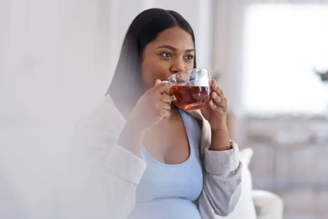 Une femme assise portant un décollecté bleu sous un pull-over gris à manches longue, tient un breuvage dans un verre qu'elle boit.