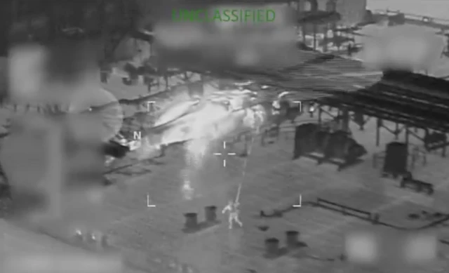 Чорно-білий кадр із тепловізор: на палубі великого судна видно людину, яка спускається на тросі з вертольота. Вертоліт зависає над палубою, його корпус і лопаті розмиті через рух. На палубі розташовані металеві конструкції та кілька великих циліндричних об’єктів. У центрі кадру є прицільні маркери, а вгорі зеленим текстом написано "UNCLASSIFIED" – "незасекречено".