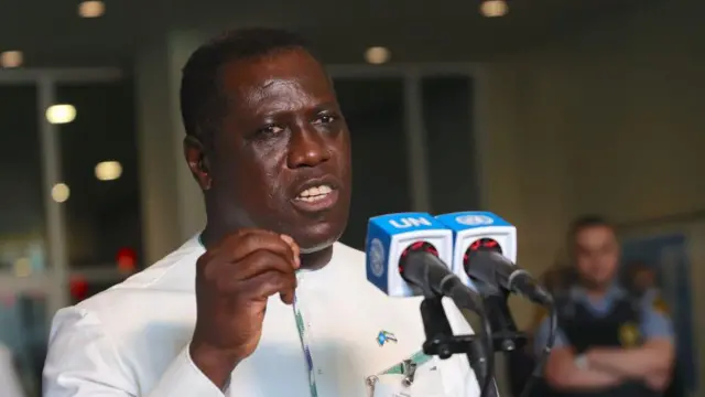 David John Francis, Waziri Mkuu wa zamani wa Sierra Leone akizungumza katika vipaza sauti viwili vyenye chapa ya Umoja wa Mataifa katika Umoja wa Mataifa mjini New York baada ya nchi yake kuchaguliwa katika Baraza la Usalama la Umoja wa Mataifa kwa muhula wa 2024-2025 Juni 6, 2023. Amevaa suti nyeupe iliyo na bendera ndogo ya nchi yake imebandikwa kifuani mwake. Kuna mlinzi kwa nyuma amesimama na mikono yake imevuka kifua chake.