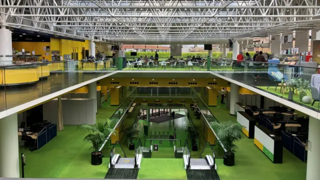The interior of a large, open-plan office building. Three floors are visible. The lower one has a modern entrance with pass barriers. Two escalators take people to the next floor, which has green flooring. The third level is a mezzanine one with yellow, green flooring, walls and desks.  The ceiling has metal struts and glass above.