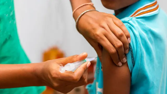 A child in a blue top sits as they receive a vaccine in their arm, from a health care professional standing next to them.