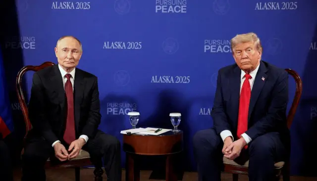 Putin e Trump sentados em frente a painel com os dizeres "Alasca 2025"