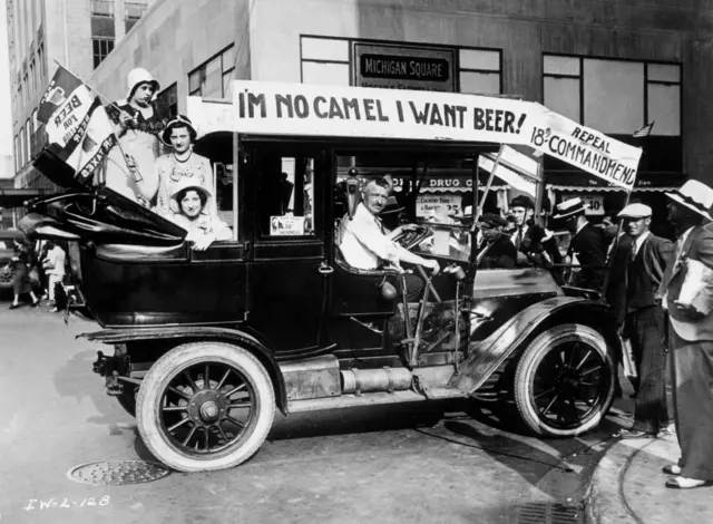 Manifestantes contra a Lei Seca, nos Estados Unidos, desfilam em um carro adornado com cartazes e bandeiras pedindo a revogação da 18ª Emenda. Um cartaz diz: "NÃO SOU CAMELO, QUERO CERVEJA!"