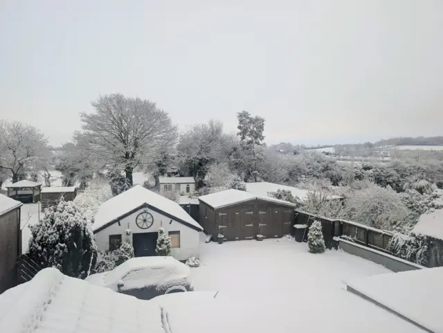 El jardín trasero de la casa está cubierto de nieve. Hay dos cobertizos pequeños. El de la izquierda es blanco, con el techo cubierto de nieve, y el de la derecha es negro, también con el techo cubierto de nieve. Un coche está estacionado frente al cobertizo blanco, cubierto de nieve. Detrás del jardín hay una valla de madera negra. Más allá de la valla hay otro jardín y un grupo de árboles altos cubiertos de nieve.