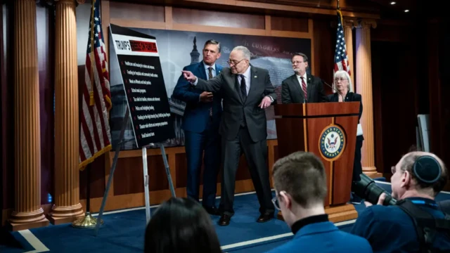 السيناتور الديمقراطي تشاك شومر، زعيم الديمقراطيين، مرتدياً بدلة رسمية سوداء، يقف على منصة في مؤتمر صحفي وأمامه لوح شارحاً وجهة نظره. وخلفه علما الولايات المتحدة وأيضاً عدد من المسؤولين