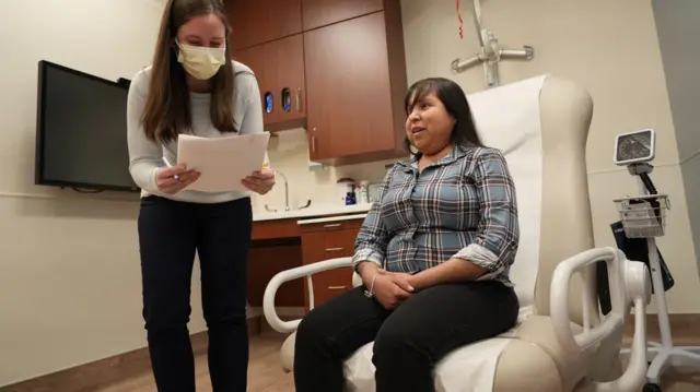 Una enfermera coordinadora de trasplantes de corazón junto a Jessica López, sentada en una silla en la habitación del hospital.