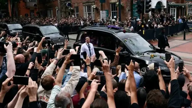 Черная машина с цветами на капоте, за ней еще несколько машин, они едут в окружении толп фанатов с поднятыми руками. В руках телефоны