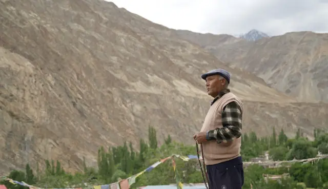 एक व्यक्ति ऊंचे पहाड़ों से घिरी घाटी में खड़ा है. उसके बाएं हाथ में प्रार्थना की माला है.