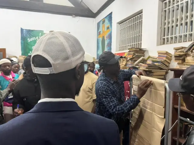 Un homme vêtu d’une chemise à carreaux bleus et d’une casquette noire, écrit sur un tableau en carton, devant lui des piles de livres sont empilés sur un tableau recouvert de papier kraft. Devant lui, on distingue des piles de livres entassés sur des étagères et des meubles, donnant à la pièce un air à la fois studieux et un peu encombré.