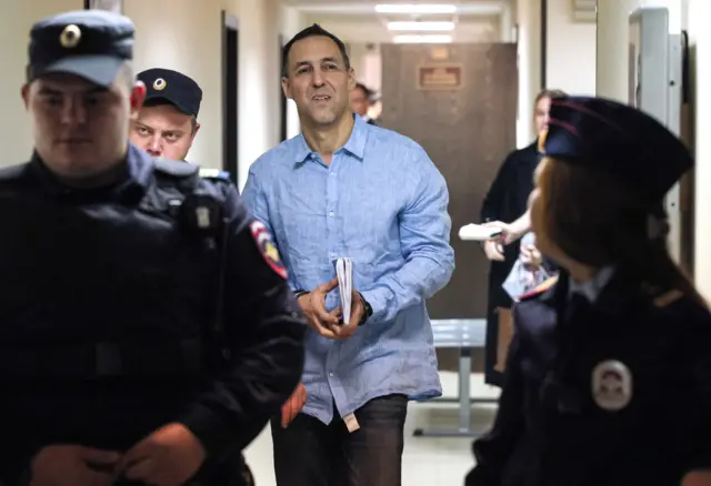 Мужчина в голубой рубашке с наручниками на запястьях и тетрадкой в руках идет с конвоирами по коридору суда в России