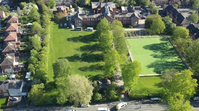 An aerial view of an ornate building with a large field in front of it as well as a playing field and trees. There are rows of houses to the left hand side of the image.