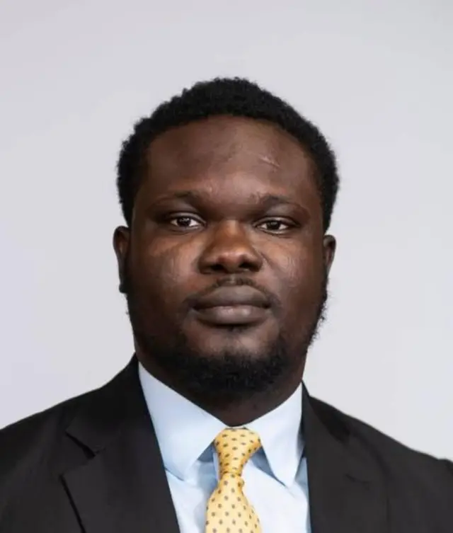 Yekinni Femi wia suit for white background dey look directly at di camera