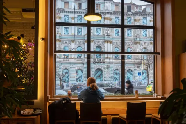 Вид зсередини кав’ярні на пошкоджену ракетним ударом будівлю колишнього універмагу. Через велике вікно видно фасад будівлі з вибитими вікнами та слідами руйнувань. На передньому плані – інтер’єр кав’ярні з дерев’яним столом, квітами та ліхтариками. 