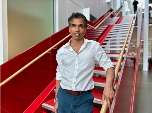 Prof Channa Jayasena stands at the bottom of a red staircase in a hospital. He has short dark hair and is wearing a white shirt with navy chinos. 