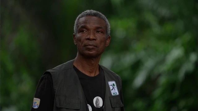 Chukwuemeka Igwe, stand for di sacred forest in Awka-Etiti. E wear an NCF badge on his gilet and a T-Shirt with photos of vultures.
