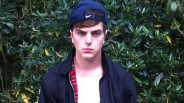 Will Adolphy de adolescente. Viste una remera blanca con una chaqueta de cremallera azul y una gorra azul oscura volteada hacia atrás. Mira con enfado a cámara 