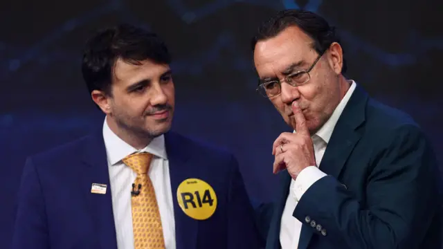 Dois homens aparecem lado a lado durante um debate. Renan Santos, à direita, de terno escuro e óculos, leva o dedo indicador à boca em um gesto de silêncio, enquanto Augusto Cury, à esquerda, de terno azul e gravata amarela, olha em sua direção. Eles estão diante de um fundo escuro iluminado em tons azulados.