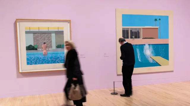 Dwóch zwiedzających muzeum przechodzi obok obrazów Davida Hockneya przedstawiających baseny.