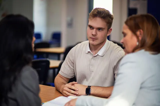 Illia listening in a tutorial. He is wearing a cream t-shirt with a collar. There are two woman sat opposite him.