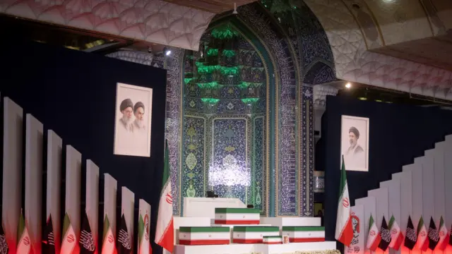 Caixões cobertos com a bandeira iraniana descansam sobre uma plataforma ladeada por bandeiras e retratos dos aiatolás