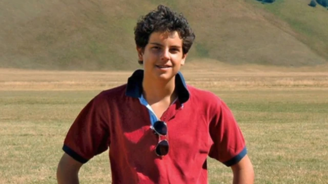 
Um garoto de cabelos cacheados e escuros, vestindo uma camisa polo vermelha, sorri para a câmera com as mãos na cintura, diante de um campo com colinas ao fundo.
