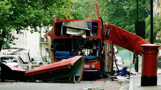 爆破されたロンドンの赤いダブルデッカーバスが、路上に止まっている。屋根が吹き飛ばされ手前の路上に落下し、車内も破壊されている
