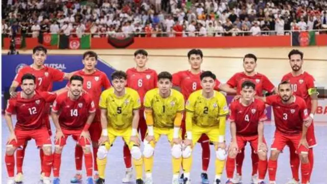 بازیکنان افغانستان قبل از بازی مقابل ایران تصویر می‌گیرند که بازیکنان با لباس‌های سرخ و دروازه‌بان با لباس‌های زرد هستند و در پس زمینه آنان تماشاچیان افغانستان که پرچم افغانستان نیز دیده می‌شود.