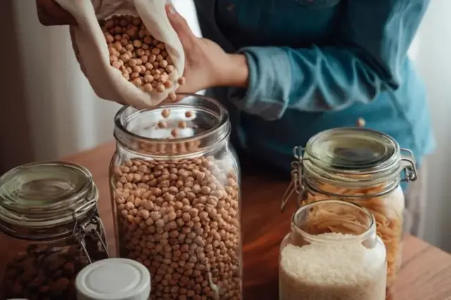 چند شیشه حبوبات روی میز، دستی که در حال ریختن نخود در یک شیشه است