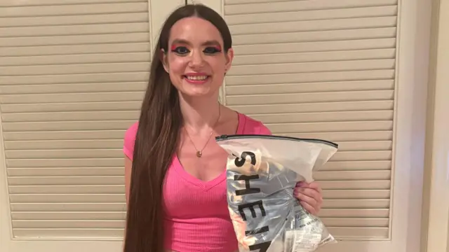 Deborah Grushkin, com os olhos fortemente maquiados, usa uma camisa rosa e segura um pacote da Shein.